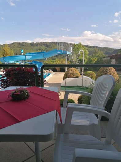 Ein gemütlicher Balkon mit einem Tisch und Stühlen. Im Hintergrund sind ein Schwimmbad und eine Rutsche zu sehen, umgeben von grünen Hügeln. | © Gemeinde Mitterberg-Sankt Martin