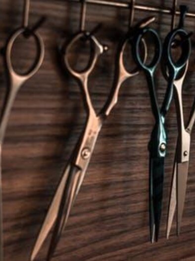 A variety of different scissors hangs on a hook. The scissors are made of metal and gleam in the light. | © Friseur Tip Top