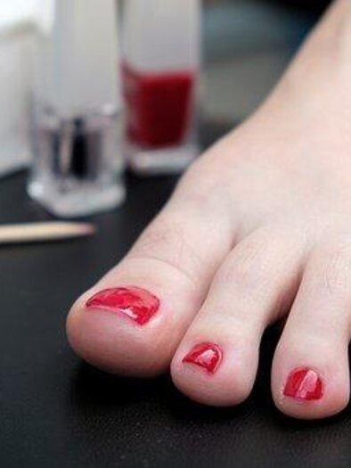 A foot with red-painted toenails is lying on a table. In the background, there are nail polish bottles and a wooden stick visible.