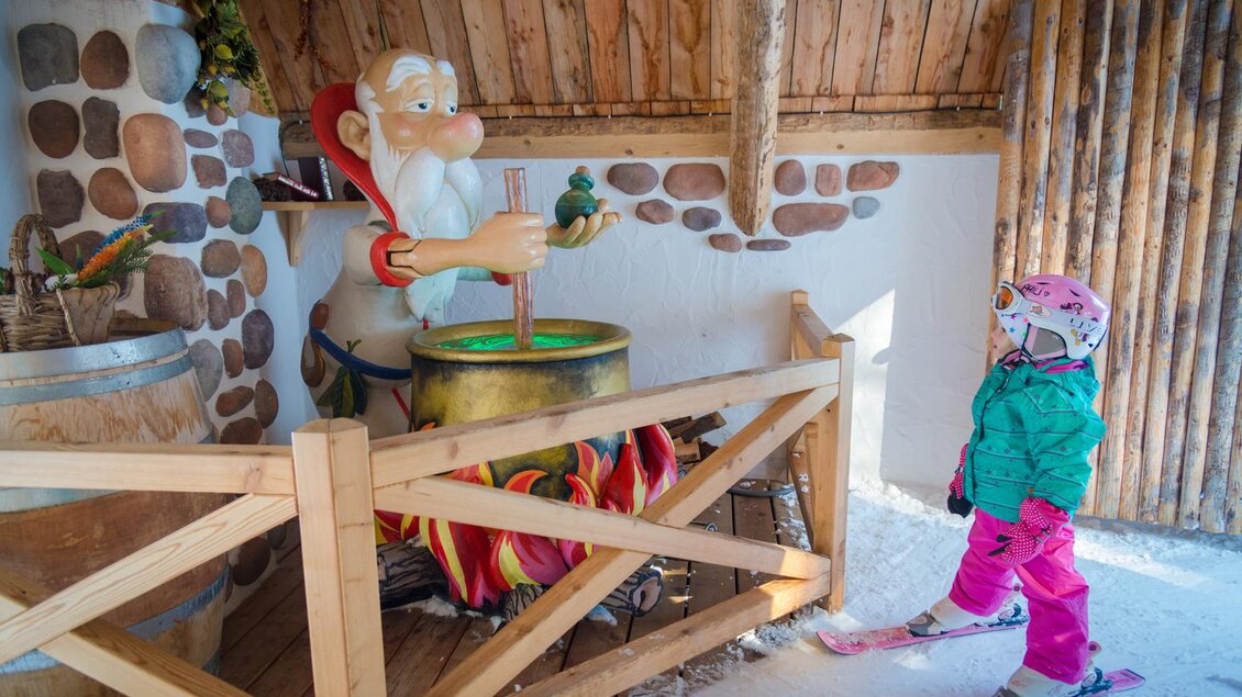 Ein kleiner Junge steht auf Skiern und schaut einen fröhlichen Weihnachtsmann an, der in einem Topf rührt. Die Szenerie ist winterlich und gemütlich, umgeben von Holzstrukturen. | © Christine Höflehner