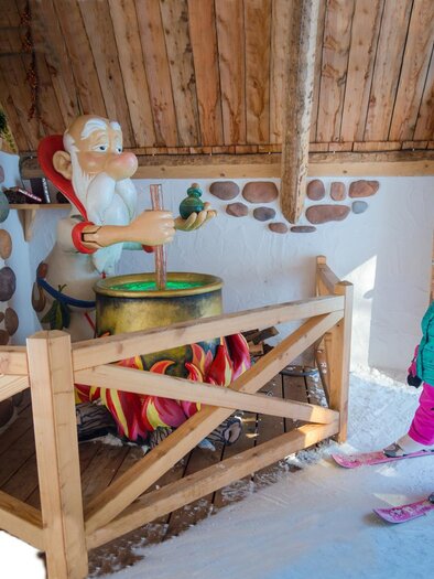 A little boy is standing on skis and looking at a cheerful Santa Claus who is stirring in a pot. The scene is wintry and cozy, surrounded by wooden structures. | © Christine Höflehner