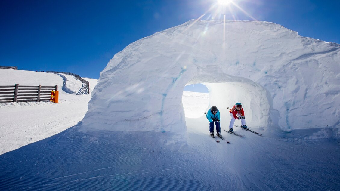 Zwei Skifahrer stehen vor einem großen Schneetunnel auf einer sonnigen Skipiste. Der klare blaue Himmel strahlt über die verschneite Landschaft. | © (c) Tom Lamm