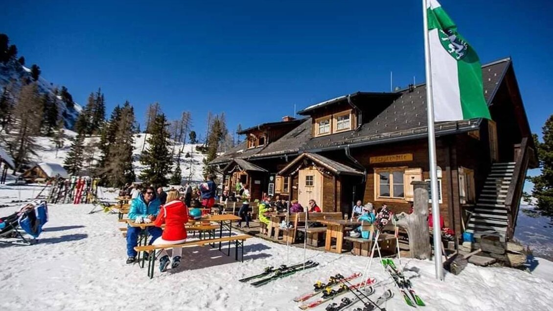 Eine gemütliche Berghütte im Schnee mit Gästen, die an Tischen sitzen. Im Vordergrund sind Skier und Bäume zu sehen, und blauer Himmel darüber. | © Galsterbergalmhütte