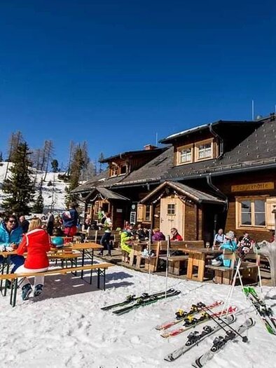 A cozy mountain cabin in the snow with guests sitting at tables. In the foreground, skis and trees can be seen, and a blue sky above. | © Galsterbergalmhütte