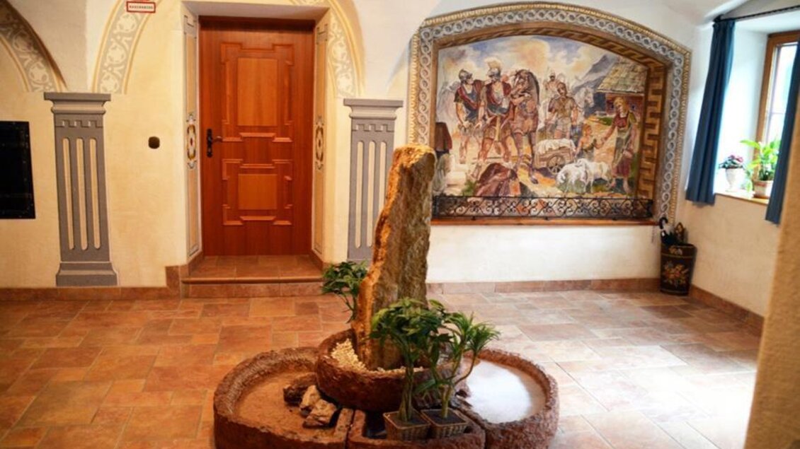 Ein einladender Eingangsbereich mit einer hölzernen Tür und einer Wandmalerei. In der Mitte befindet sich ein schöner Brunnen mit Pflanzen. | © Christian Tritscher