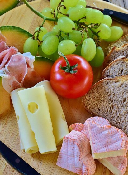 Ein Holzbrett mit verschiedenen Lebensmitteln: Melonenscheiben, Trauben, Schinken, Käse, Tomate und Brotscheiben. Dazu liegen zwei Messer auf dem Brett. | © Symbolfoto Pixabay