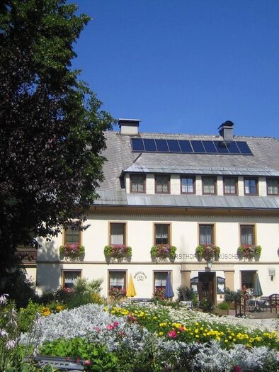 Ein charmantes Gasthaus mit blühenden Gärten und einem blauen Himmel. Die Fassade ist freundlich und einladend, mit vielen Pflanzen und Tischen im Freien. | © Gasthof Rüscher