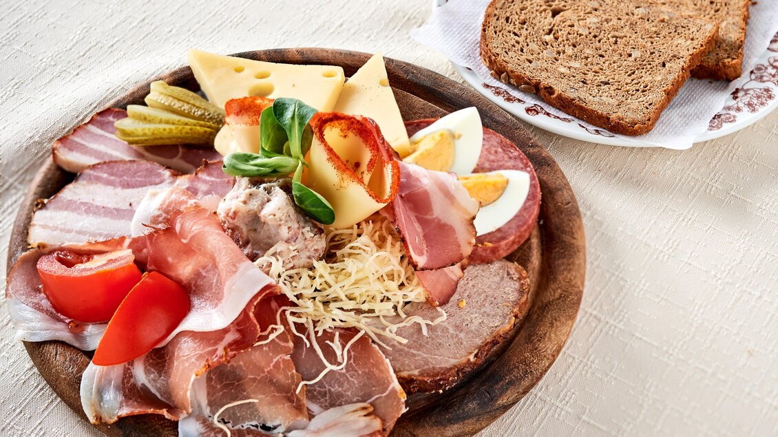 Ein rustikeller Teller mit verschiedenen Wurst- und Käsesorten, garniert mit frischem Gemüse. Daneben liegt eine Scheibe dunkles Brot. | © Gasthof Stockerwirt
