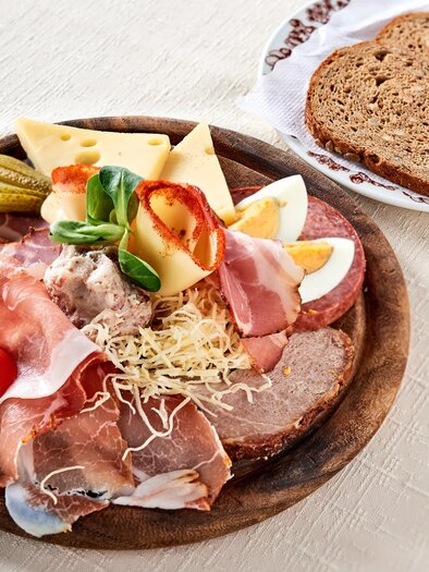 Ein rustikeller Teller mit verschiedenen Wurst- und Käsesorten, garniert mit frischem Gemüse. Daneben liegt eine Scheibe dunkles Brot. | © Gasthof Stockerwirt