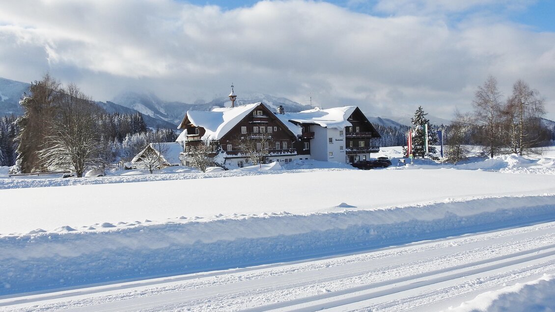 Ein schönes, verschneites Landschaftsbild mit einem traditionellen Gebäude im Vordergrund. Im Hintergrund sind Berge und ein blauer Himmel zu sehen. | © Gasthof Stockerwirt