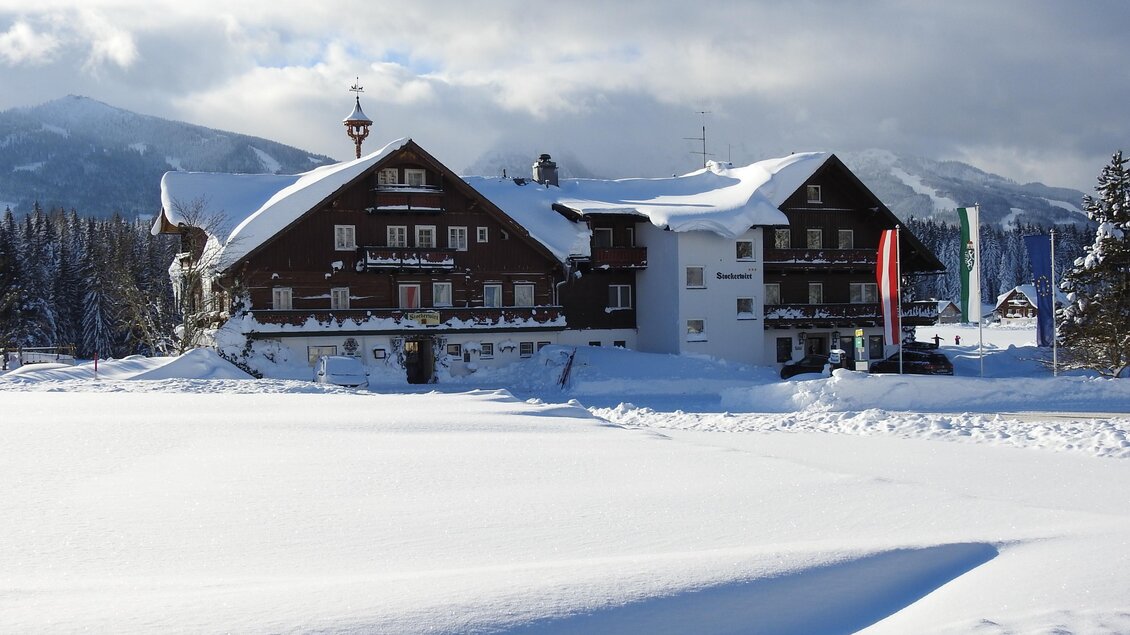 Ein hübsches Gebäude im Schnee, umgeben von einer winterlichen Landschaft. Die Wolken sind klar und die Berge sind im Hintergrund sichtbar. | © Gasthof Stockerwirt