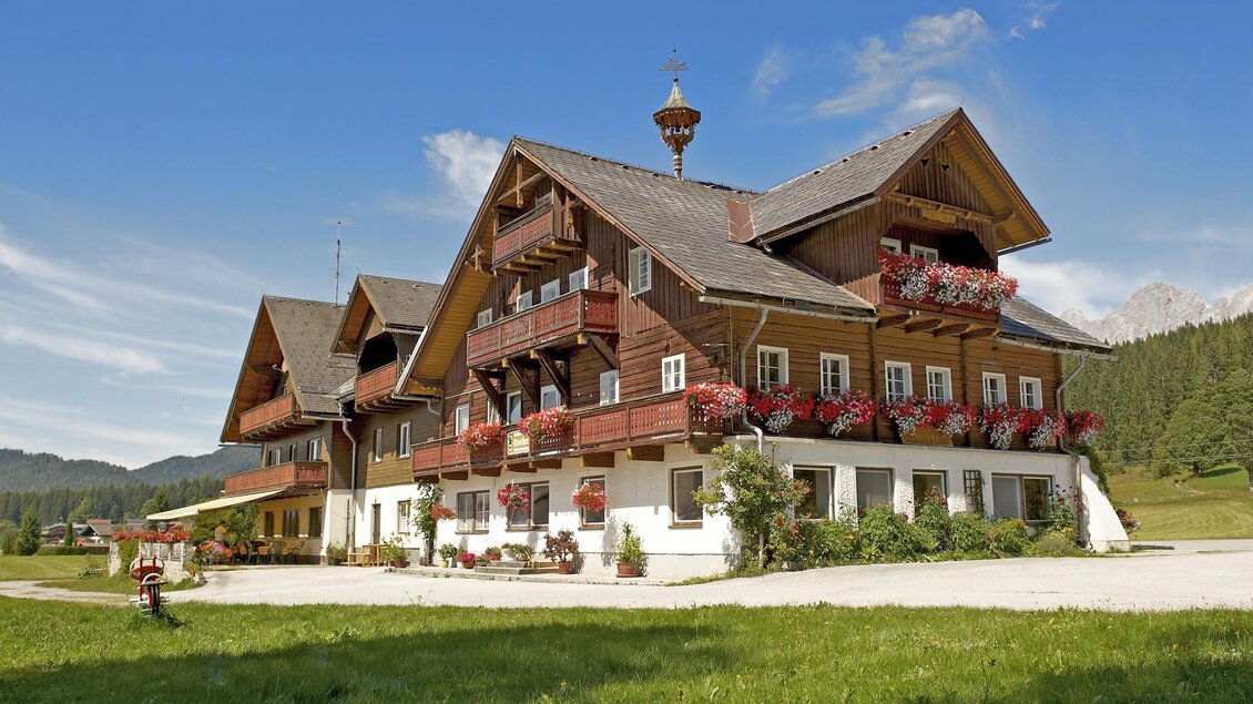 Ein schönes, traditionelles Chalet mit Balkonen und blühenden Blumen. Umgeben von einer malerischen Landschaft und einem klaren blauen Himmel. | © Gasthof Stockerwirt