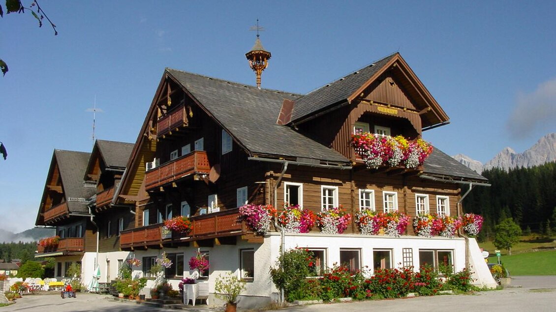 Ein traditionelles, mehrstöckiges Holzhaus mit schönen Blumen auf den Balkonen. Im Hintergrund sind sanfte Hügel und Bäume zu sehen. | © Gasthof Stockerwirt