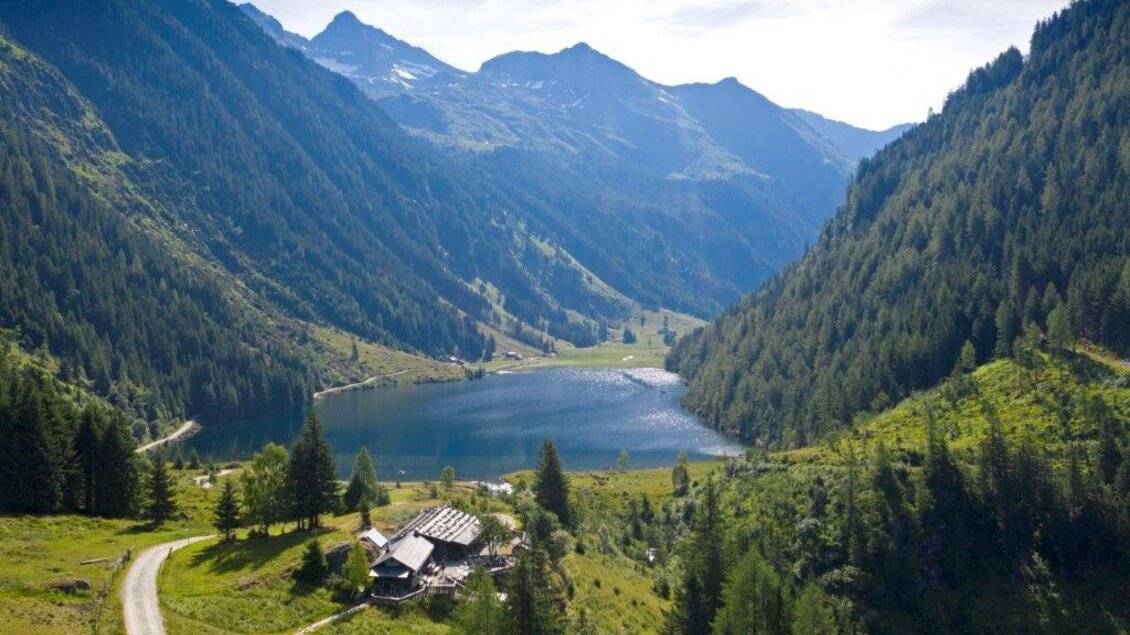 Eine malerische Landschaft mit Bergen und einem klaren See. Im Vordergrund ist ein charmantes Holzhaus zu sehen, umgeben von üppigem Grün. | © Herbert Raffalt