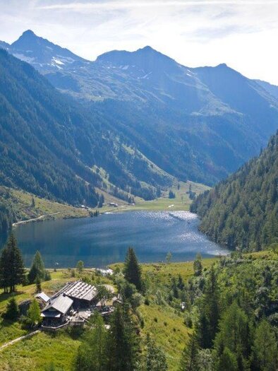 A picturesque landscape with mountains and a clear lake. In the foreground, a charming wooden house can be seen, surrounded by lush greenery. | © Herbert Raffalt