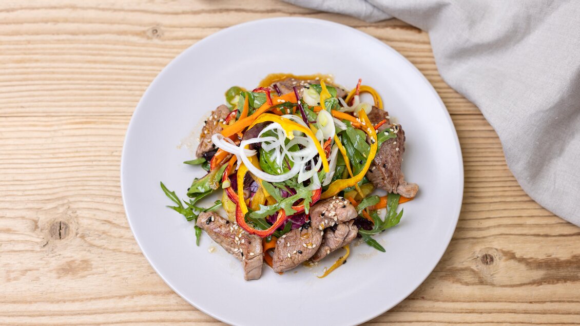 Ein Teller mit Fleisch und buntem Gemüse, garniert mit Frühlingszwiebeln und Sesam. Die Speise ist auf einem Holztisch angerichtet. | © Martin Huber