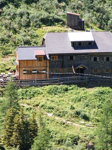 A large house made of stone and wood, surrounded by green trees and bushes.  
On the porch, there are some seating options and there is a beautiful view of nature. | © Gollinghütte
