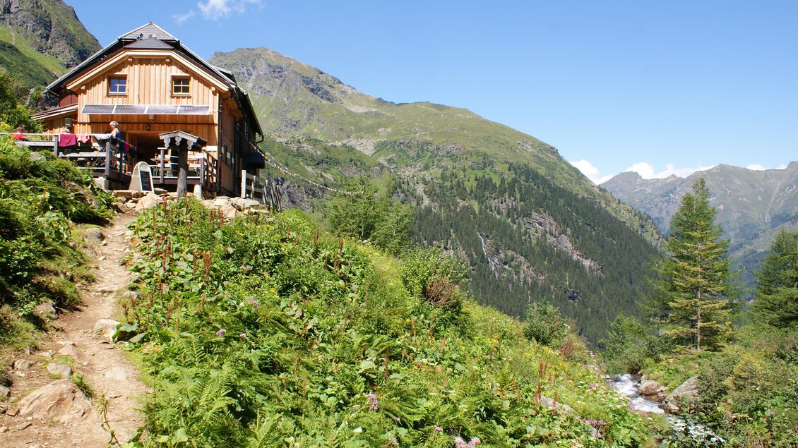 Ein altes Holzhaus steht malerisch auf einem Hügel umgeben von grünen Wiesen. Im Hintergrund sind majestätische Berge und ein klarer blauer Himmel zu sehen. | © Gollinghütte