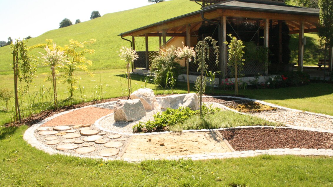 Ein schöner Garten mit verschiedenen Pflanzen und dekorativen Steinen. Im Hintergrund ist ein modernes Gebäude und sanfte Hügel zu sehen. | © Infobüro Gröbming