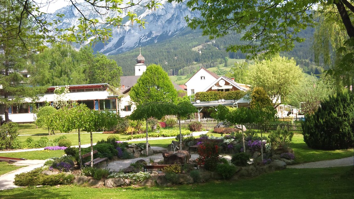 Ein gepflegter Garten mit bunten Blumen und Sträuchern, umgeben von Bäumen. Im Hintergrund sind die Berge und ein kleines Gebäude mit einer Kirchturm zu sehen. | © Infobüro Gröbming