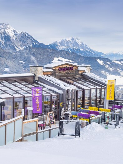 Exterior view of the Gumpenbar at Hauser Kaibling with glass front, panoramic terrace, and view of the snow-covered mountains. | © Gumpenbar