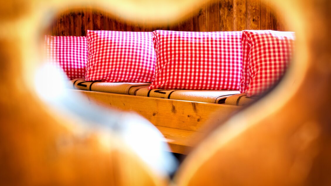 Ein gemütliches Holzinterieur mit roten karierten Kissen. Die Sicht ist durch ein herzförmiges Loch im Holz gerahmt. | © René Eduard Perhab