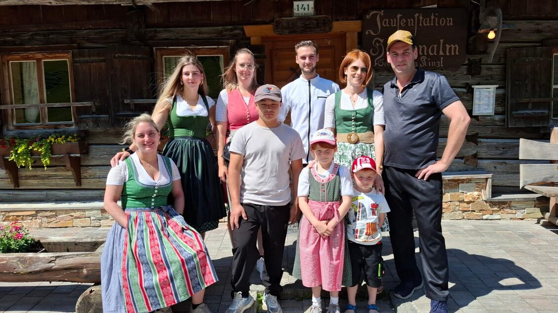 Eine fröhliche Gruppe von Menschen in traditionellen Trachten steht vor einem rustikalen Holzgebäude. Die Sonne scheint, und die Stimmung wirkt entspannt und freundlich. | © Martin Heller, Hansenalm