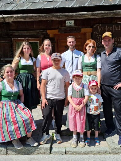 A cheerful group of people in traditional costumes stands in front of a rustic wooden building. The sun is shining, and the atmosphere feels relaxed and friendly. | © Martin Heller, Hansenalm