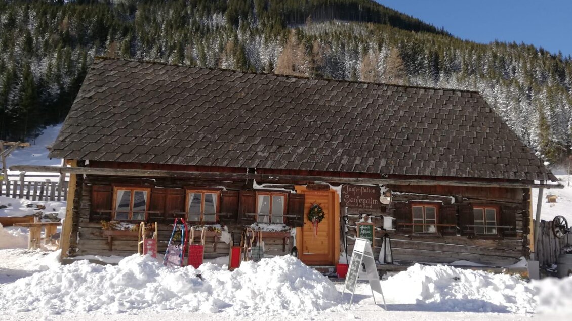 Eine gemütliche Holzhütte im Schnee, umgeben von verschneiten Bergen. Vor der Hütte stehen verschiedene Schlitten und ein Schild. | © Haiger Stefanie