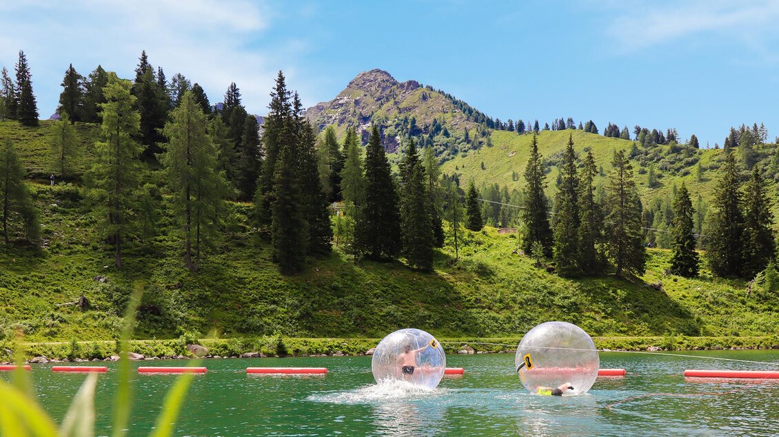 Zwei Personen in durchsichtigen Wasserbällen schwimmen auf einem grünen See. Im Hintergrund erstrecken sich Wälder und Hügel unter einem blauen Himmel.