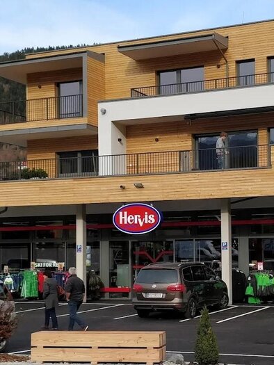 A modern store named Hervis with a wooden facade. Customers are standing in front of the entrance and looking at sports clothing. | © Hervis Sport Schladming