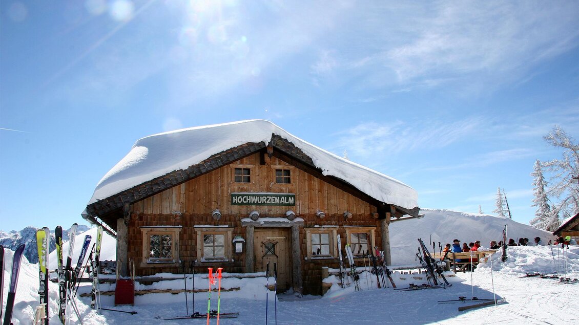 Eine gemütliche Berghütte mit einer schneebedeckten Dach. Im Vordergrund stehen Skier und die Sonne scheint klar am blauen Himmel. | © Hochwurzenalm