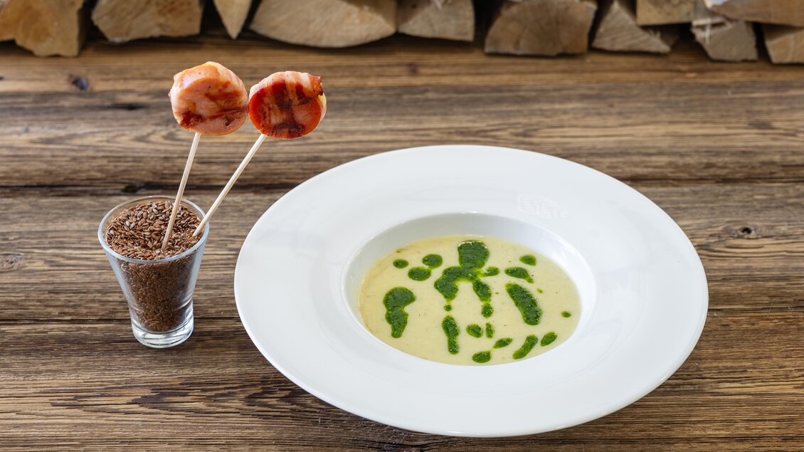 Eine cremige Suppe mit grünen Kräuterflecken auf einem weißen Teller. Daneben befinden sich zwei Spießchen mit Fleisch und ein Glas mit Gewürzen. | © Martin Huber