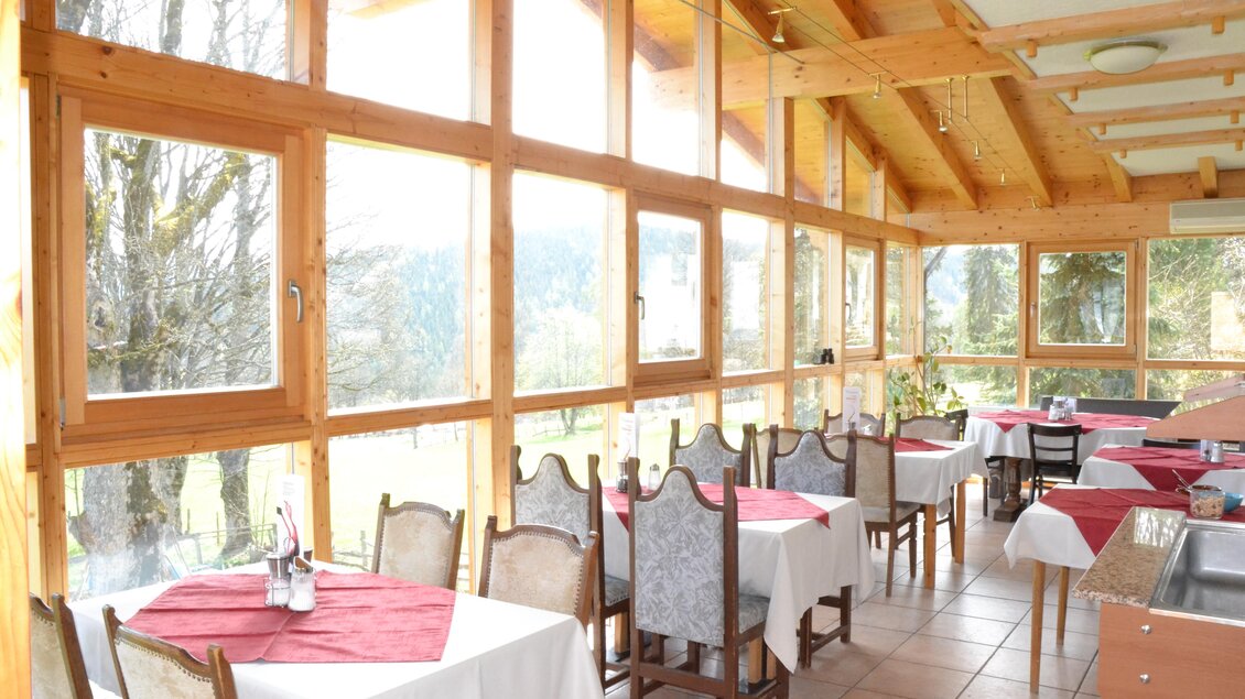 Ein helles Restaurant mit großen Fenstern und Holzverkleidung. Die Tische sind mit roten Tischdecken gedeckt und bieten Blick auf die Natur. | © Hofweyer Stube