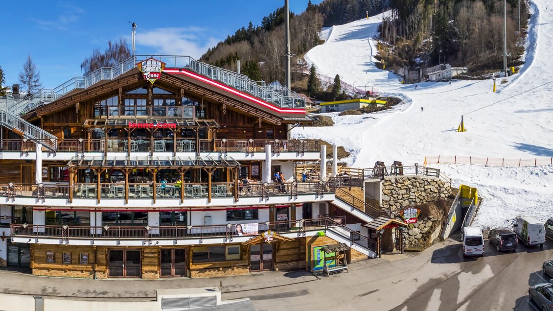 Ein alpines Skigebiet mit einem rustikalen Restaurant und vielen Autos auf dem Parkplatz. Im Hintergrund sieht man verschneite Pisten und Wälder. | © AIR-Media.at