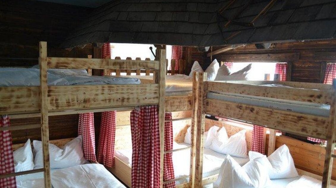 Ein gemütlicher Schlafraum mit Etagenbetten aus Holz. Die Fenster sind mit rot karierten Vorhängen geschmückt und es gibt mehrere Kopfkissen auf den Betten. | © Hoo-Ruck Alm