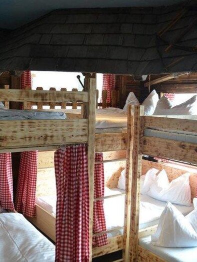 A cozy sleeping room with wooden bunk beds. The windows are adorned with red plaid curtains, and there are several pillows on the beds. | © Hoo-Ruck Alm