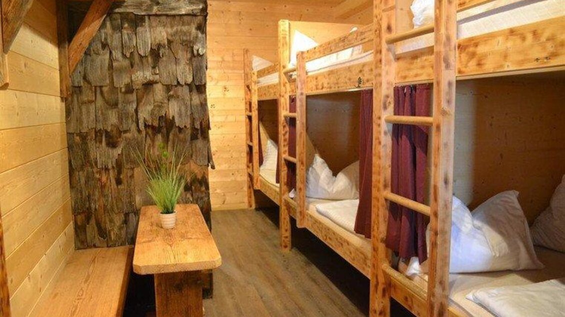 Ein gemütlicher Schlafraum mit Etagenbetten aus Holz und einer kleinen Sitzecke. Die Wände sind aus natürlichen Materialien, und es gibt einen Tisch mit einer Pflanze. | © Hoo-Ruck Alm