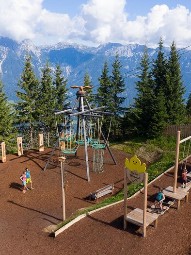 Ein Spielplatz in den Bergen mit einer Seilbahn im Hintergrund. Umgeben von Bäumen und einer schönen Berglandschaft. | © Josh Absenger