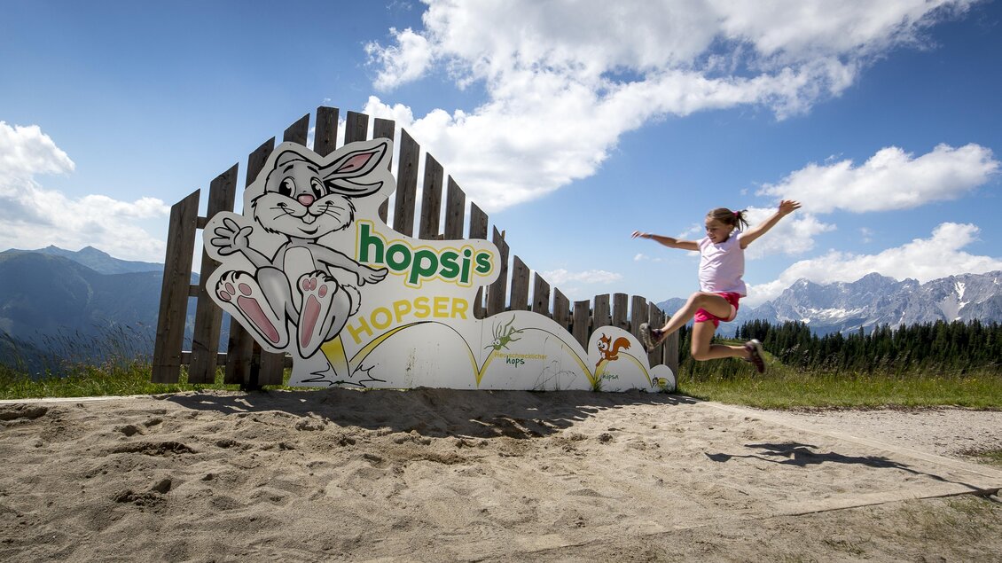 Ein Mädchen springt vor einem bunten Schild von Hopsi. Im Hintergrund sind Berge und ein blauer Himmel zu sehen. | © Tom Lamm