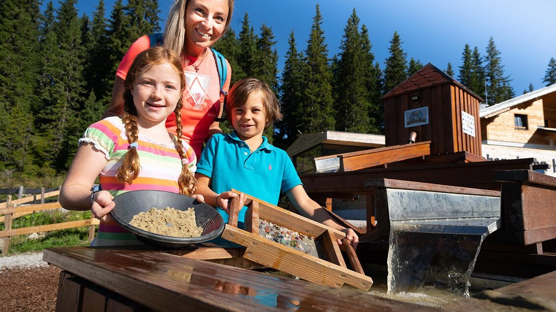 Eine Mutter steht mit ihren zwei Kindern an einem Wasserspielplatz. Die Kinder benutzen Werkzeuge, um Sand oder Kies zu durchsieben, umgeben von Bäumen und blauen Himmel. | © Josh Absenger