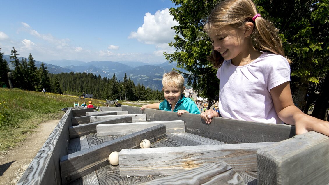 Zwei Kinder spielen mit Kugeln in einem Holzspielgerät im Freien. Im Hintergrund sind Berge und ein blauer Himmel mit ein paar Wolken zu sehen. | © Tom Lamm