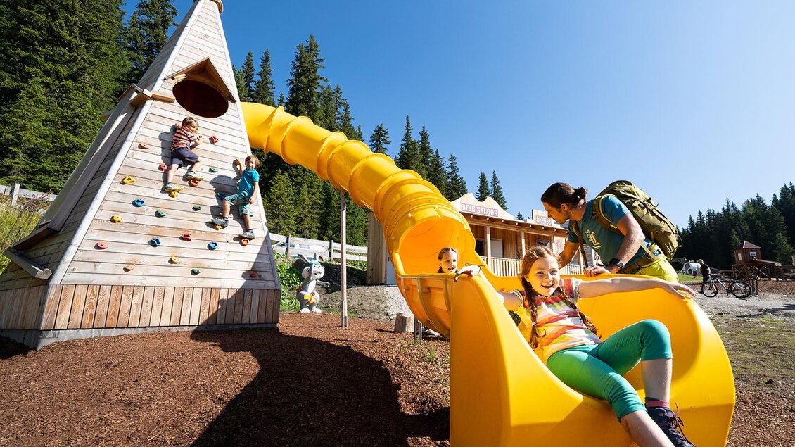 Ein Spielplatz mit einer Kletterwand und einer langen Rutsche. Kinder spielen fröhlich in der Natur unter blauem Himmel. | © Josh Absenger