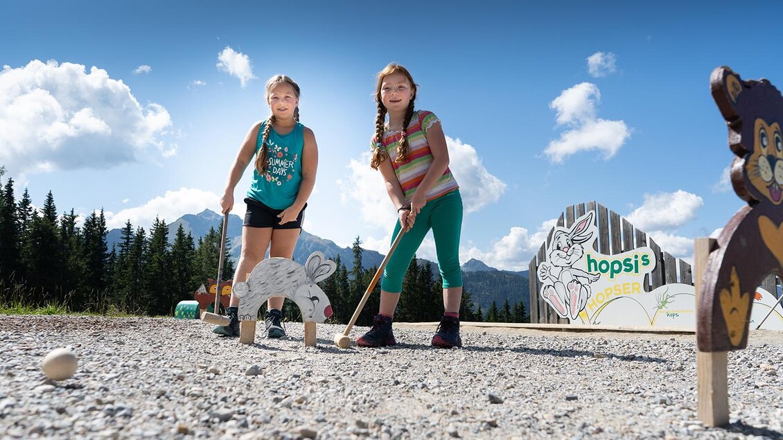 Zwei Mädchen spielen Minigolf im Freien. Im Hintergrund sind Berge und eine sonnige Himmelslandschaft sichtbar. | © Josh Absenger