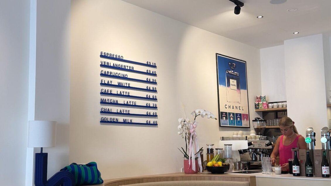Ein modernes Café mit hellen Wänden und minimalistischer Einrichtung. An der Wand hängt ein Bild, daneben steht ein Mitarbeiter an der Theke. | © Hufschmiede