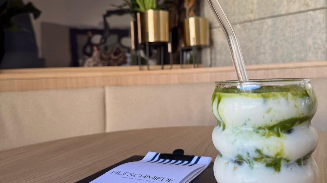 Ein farbenfroher Drink in einem dekorativen Glas steht auf einem Holztisch. Im Hintergrund sind Pflanzen und eine Speisekarte sichtbar. | © Hufschmiede