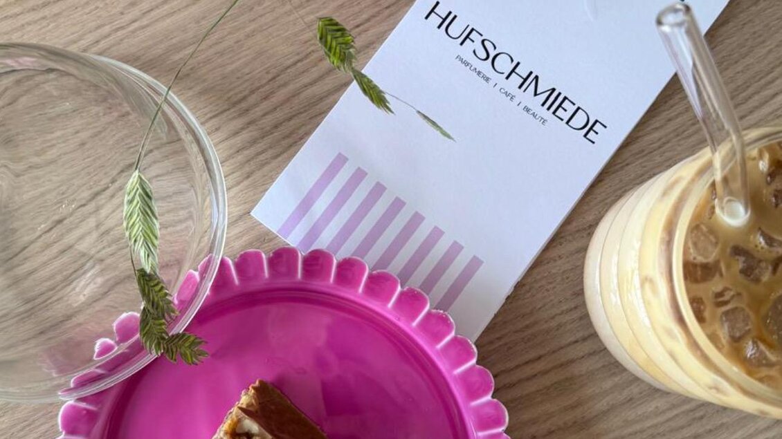 Ein Stück Kuchen auf einem pinken Teller neben einem Glas Eiskaffee. Im Hintergrund sind Blumen und eine Karte zu sehen. | © Hufschmiede
