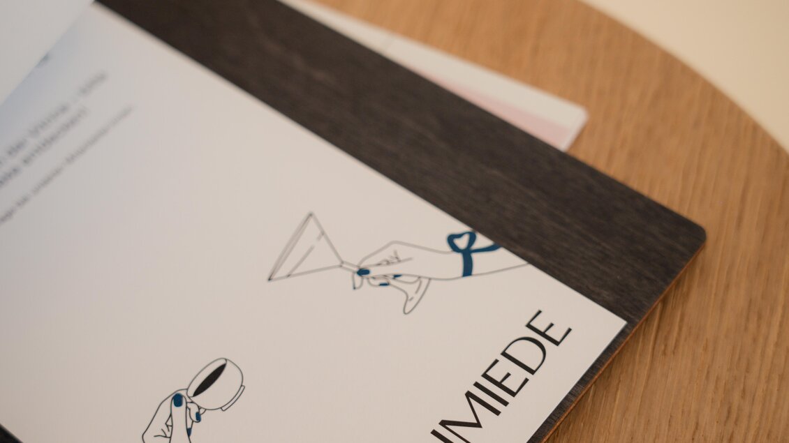 Ein geöffnetes Menü mit dem Titel "HUFSCHMIEDE". Auf der Seite sind einfache Illustrationen von Händen, die ein Glas und eine Flasche halten. | © Hufschmiede