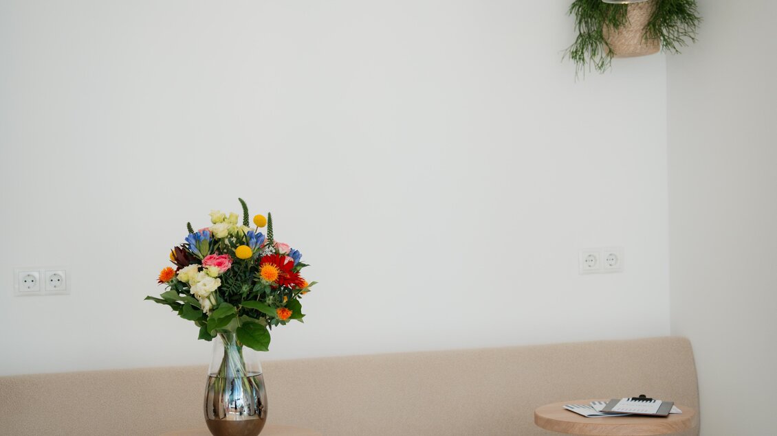 Ein modischer Wartebereich mit einer beigen Bank und modernen Tischen. Auf einem Tisch steht eine bunte Blumenvasenarrangement. | © Hufschmiede