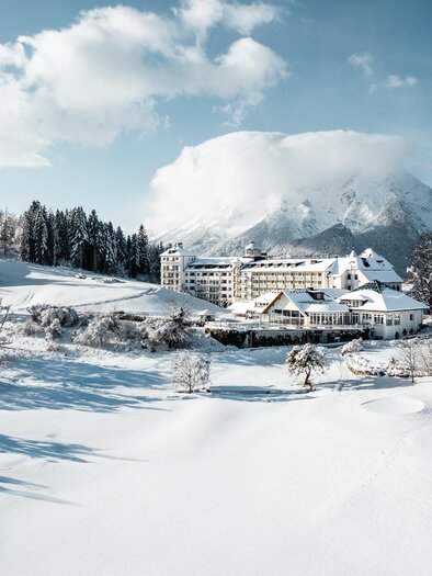 A snowy landscape with a large mountain in the background. In the foreground stands a charming hotel, surrounded by snow-covered trees. | © Thomas Sattler